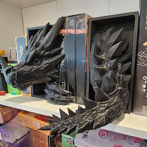 Op de afbeelding: Zwarte draken boekensteunen. De boekensteunen zijn gemaakt van een donkergrijs materiaal en zijn ontworpen om op een draak te lijken. De kop en staart zijn aan weerszijden van een rij boeken geplaatst.