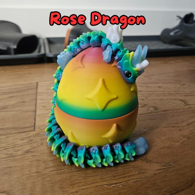 May include: A 3D printed, multi-colored dragon figurine curled around a rainbow egg with stars. The dragon has a white horn and blue wings. The text "Rose Dragon" is written in red above the figurine.