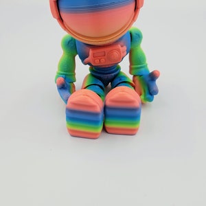 May include: A rainbow-colored astronaut figurine with a helmet and a small radio on its chest. The figurine is sitting with its legs crossed.
