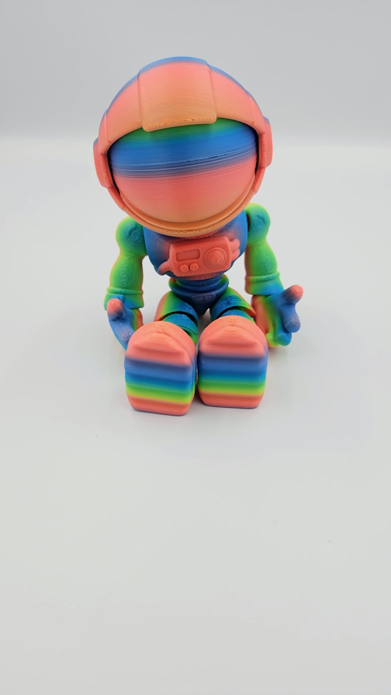 May include: A rainbow-colored astronaut figurine with a helmet and a small radio on its chest. The figurine is sitting with its legs crossed.