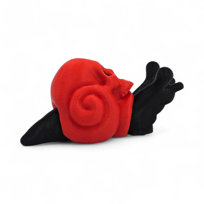 May include: A red skull-shaped snail ornament with a spiraled shell and black body. The snail's body is black and the shell is a vibrant red. The ornament is set against a white background.