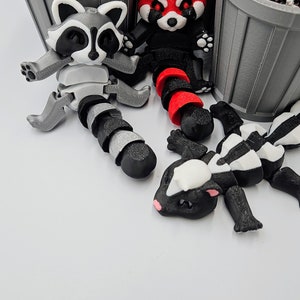 May include: A collection of articulated animal figures, including a raccoon, red panda, and skunk, with segmented bodies. The figures are black, white, and gray, with red accents. Gray trash cans filled with colorful objects are in the background.