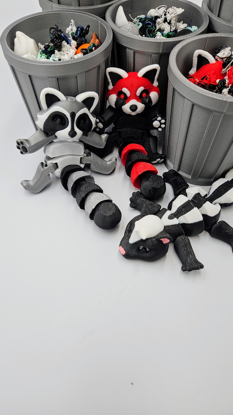 May include: A collection of articulated animal figures, including a raccoon, red panda, and skunk, with segmented bodies. The figures are black, white, and gray, with red accents. Gray trash cans filled with colorful objects are in the background.
