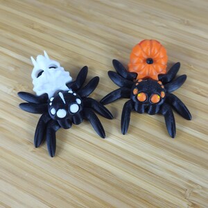 Puede incluir: Dos ara&ntilde;as negras con detalles blancos y naranjas. Una ara&ntilde;a tiene una cabeza blanca con forma de calavera y la otra tiene una cabeza naranja con forma de calabaza.