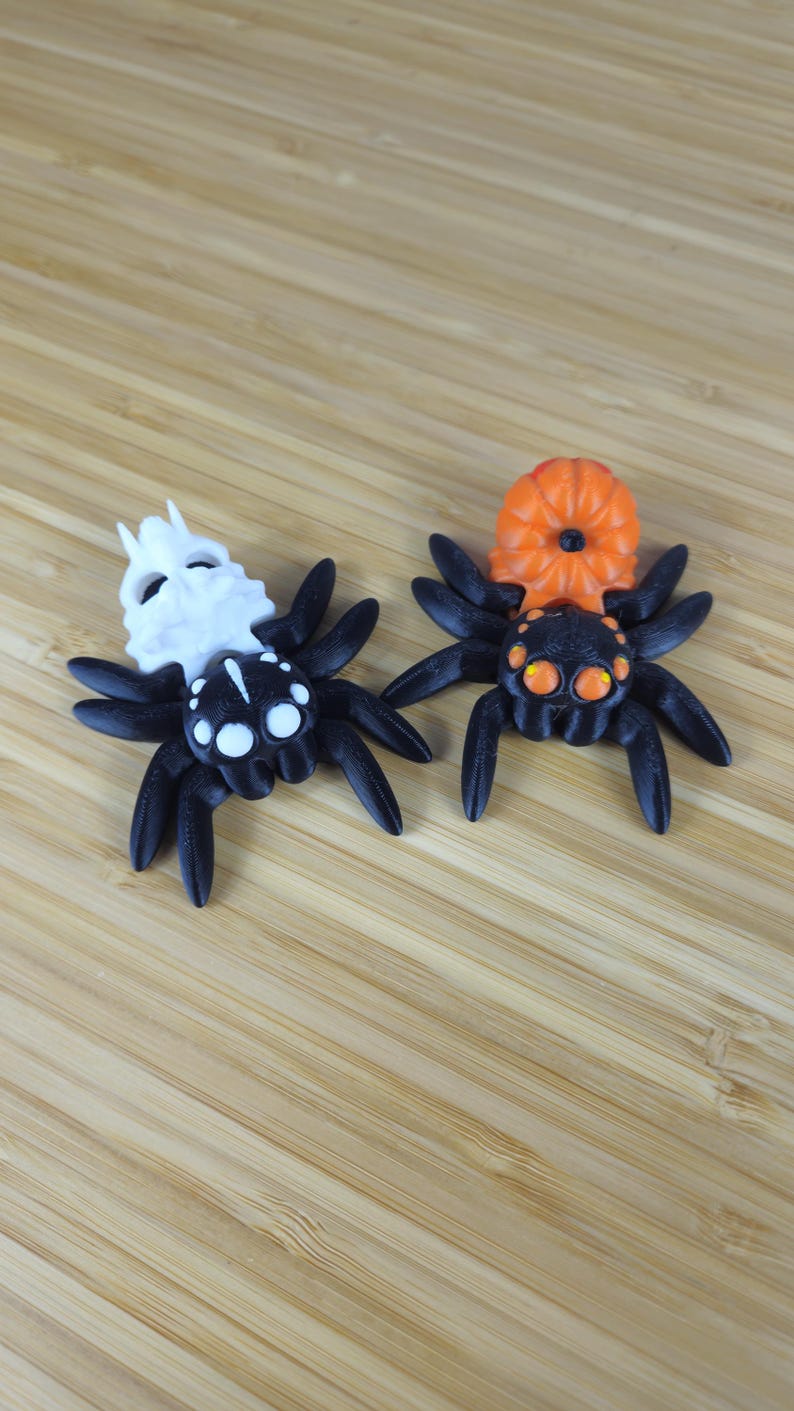 Puede incluir: Dos ara&ntilde;as negras con detalles blancos y naranjas. Una ara&ntilde;a tiene una cabeza blanca con forma de calavera y la otra tiene una cabeza naranja con forma de calabaza.