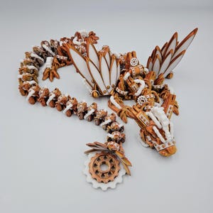 May include: A white and copper-coloured 3D printed dragon necklace with intricate details. The dragon is curled up with its wings spread out. The necklace is made of a series of connected gears and other mechanical elements.
