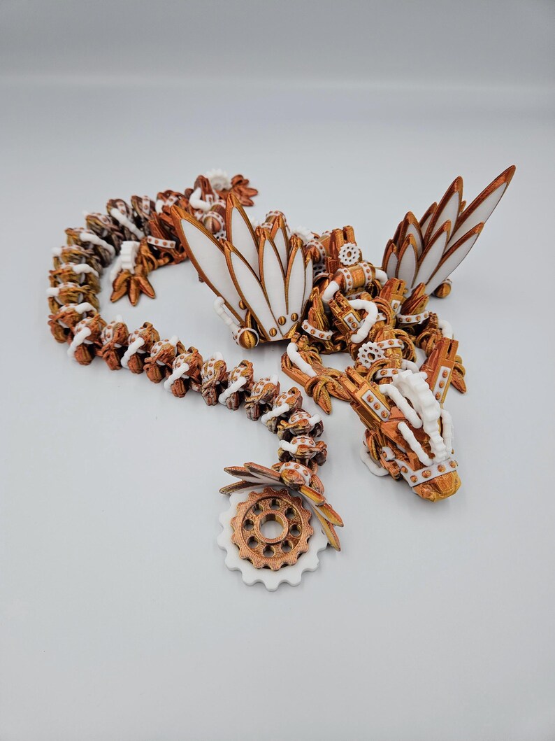 May include: A white and copper-coloured 3D printed dragon necklace with intricate details. The dragon is curled up with its wings spread out. The necklace is made of a series of connected gears and other mechanical elements.