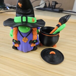 Puede incluir: Una figura de bruja impresa en 3D con un sombrero negro, detalles verdes y cabello naranja. Junto a ella hay un caldero negro con una escoba y una tapa. La bruja lleva un vestido morado y tiene detalles de ara&ntilde;as naranjas en su sombrero.
