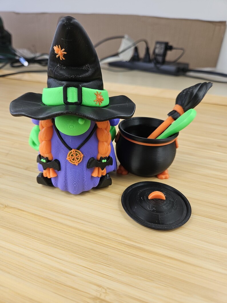 Puede incluir: Una figura de bruja impresa en 3D con un sombrero negro, detalles verdes y cabello naranja. Junto a ella hay un caldero negro con una escoba y una tapa. La bruja lleva un vestido morado y tiene detalles de ara&ntilde;as naranjas en su sombrero.