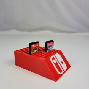 Może przedstawiać: Czerwony plastikowy stojak na kartridże do gier Nintendo Switch z miejscem na 20 kartridży. Widoczne są dwa kartridże, jeden do gry The Legend of Zelda: Breath of the Wild, a drugi do gry Super Mario Odyssey. Stojak ma białe logo Nintendo Switch z boku.