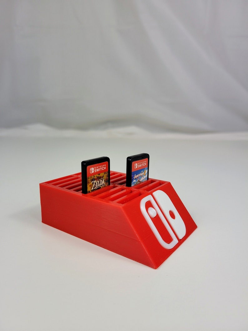 Może przedstawiać: Czerwony plastikowy stojak na kartridże do gier Nintendo Switch z miejscem na 20 kartridży. Widoczne są dwa kartridże, jeden do gry The Legend of Zelda: Breath of the Wild, a drugi do gry Super Mario Odyssey. Stojak ma białe logo Nintendo Switch z boku.