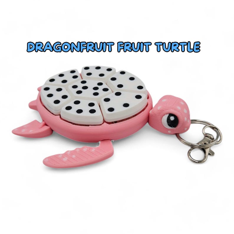 May include: A pink and white turtle-shaped keychain with a dragonfruit design. The turtle's shell is made of multiple pieces that can be moved around. The keychain has a silver metal ring.