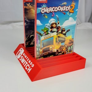 May include: Red plastic stand for Nintendo Switch game cartridges. The stand has a slot for 10 cartridges and features the Nintendo Switch logo. Two game cartridges are displayed in the stand, one for Overcooked! 2 and one for another game.