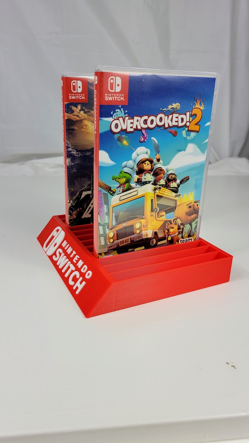 May include: Red plastic stand for Nintendo Switch game cartridges. The stand has a slot for 10 cartridges and features the Nintendo Switch logo. Two game cartridges are displayed in the stand, one for Overcooked! 2 and one for another game.