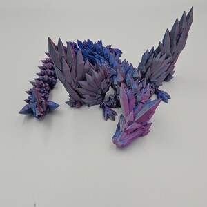 May include: A 3D printed dragon figurine with a crystal-like texture. The dragon is in a curled up position with its wings spread out. The figurine is made of a purple and blue iridescent plastic.