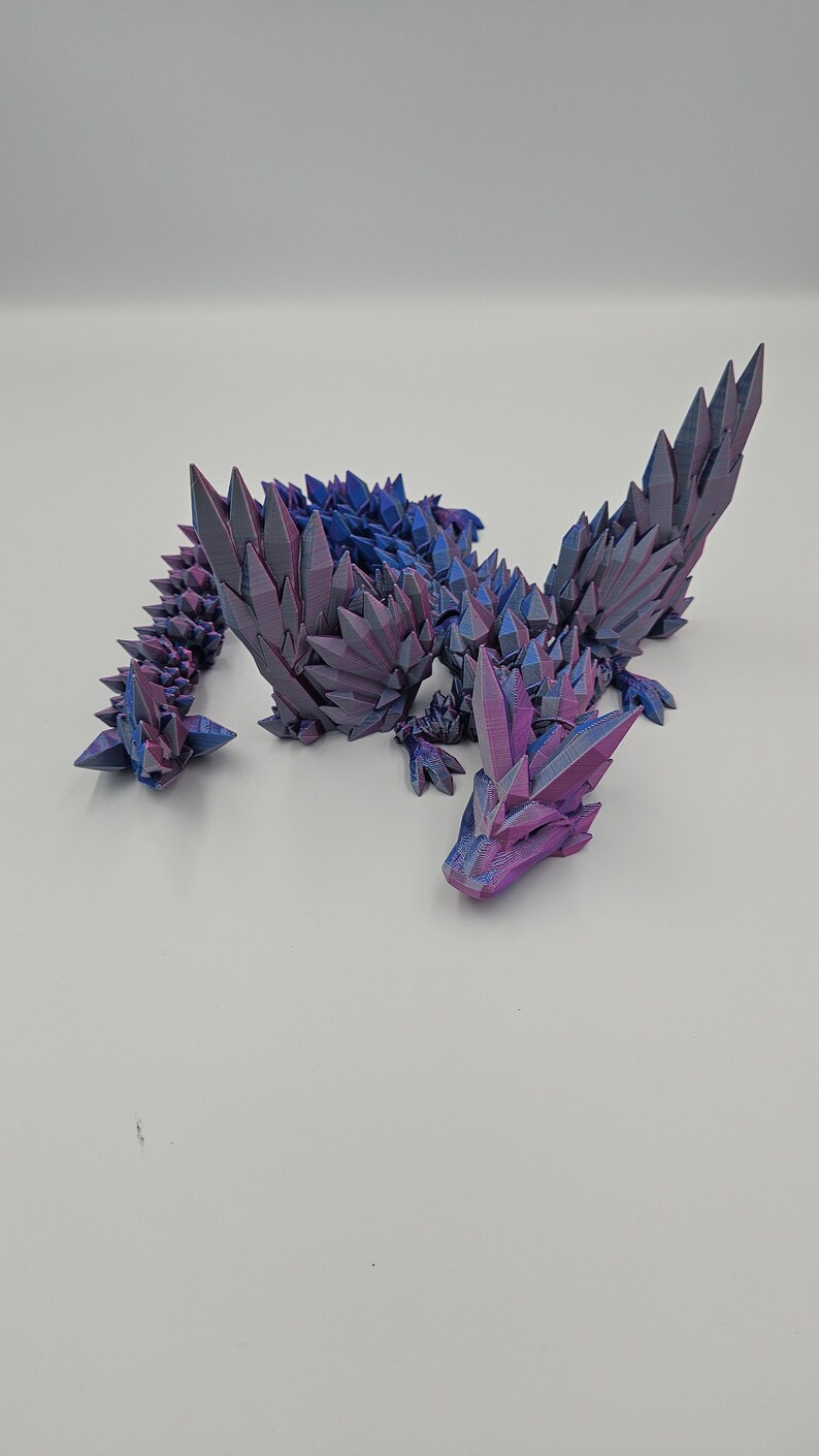 May include: A 3D printed dragon figurine with a crystal-like texture. The dragon is in a curled up position with its wings spread out. The figurine is made of a purple and blue iridescent plastic.