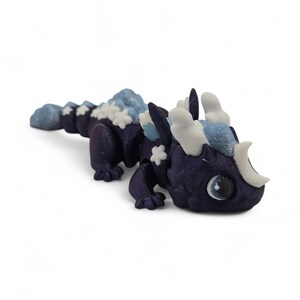 May include: A small, dark purple dragon figurine with light blue and white accents. The dragon has large, round eyes and snowflake-shaped details along its back. The figurine is a fantasy-themed collectible.