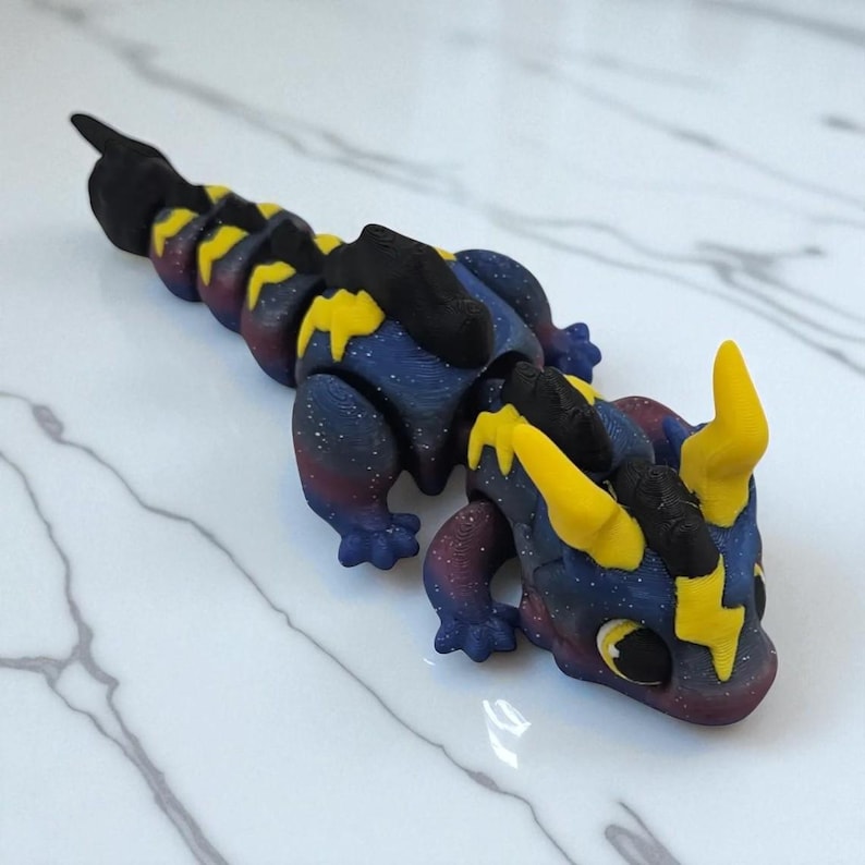 K&ouml;nnte beinhalten: Handgefertigte, bewegliche Drachenfigur. Der Drache hat einen K&ouml;rper im Galaxien-Design mit blauen, lila und wei&szlig;en Sprenkeln. Er hat gelbe Blitzakzente, H&ouml;rner und einen schwarzen R&uuml;cken. Der Drache liegt auf einer wei&szlig;en Marmoroberfl&auml;che.