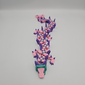 May include: A 3D-printed articulated dragon toy in pink, purple, and teal. The dragon has a long, segmented body with spikes and a detailed head. The toy is designed to be flexible and poseable.