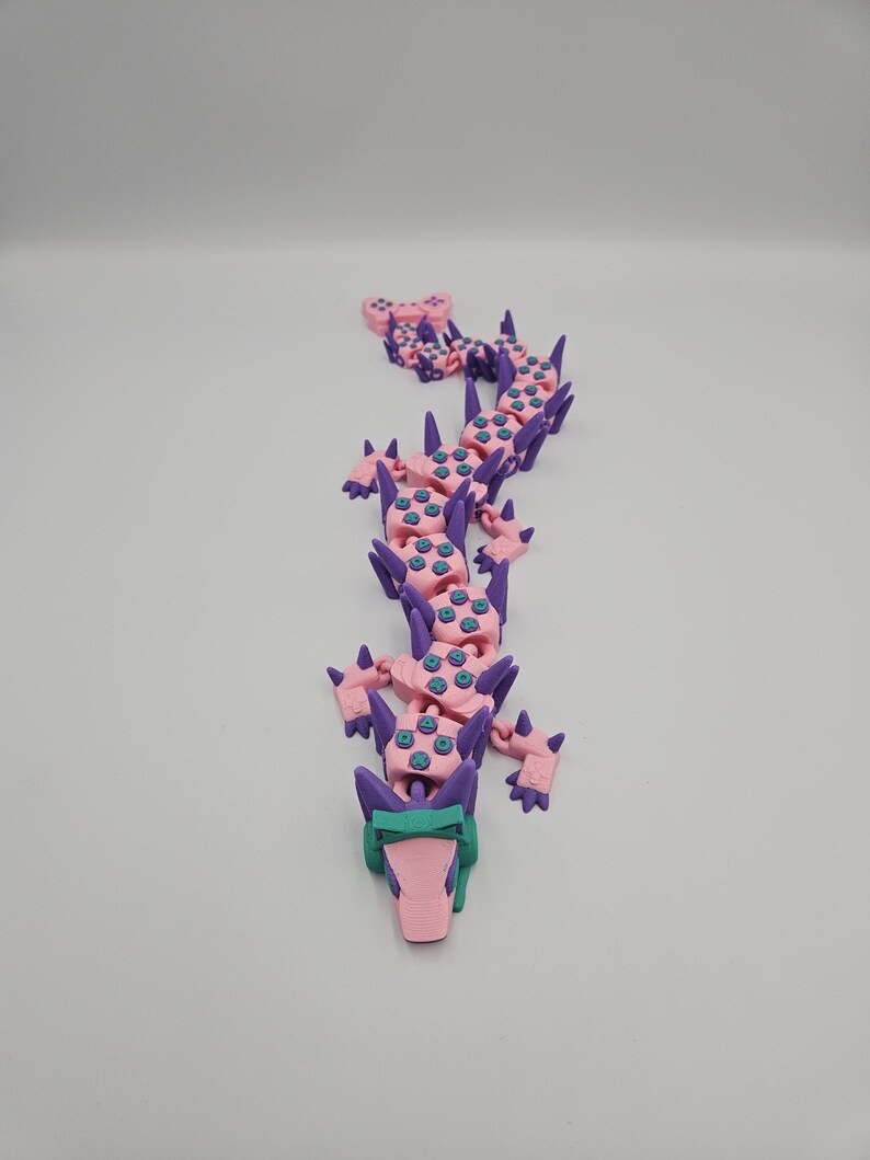 May include: A 3D-printed articulated dragon toy in pink, purple, and teal. The dragon has a long, segmented body with spikes and a detailed head. The toy is designed to be flexible and poseable.