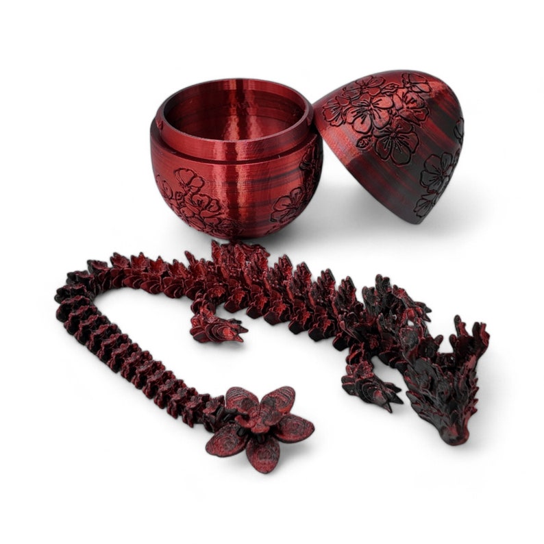 Pu&ograve; includere: Un drago rosso stampato in 3D con un motivo floreale e un contenitore a forma di uovo abbinato. Il drago &egrave; arrotolato su se stesso con la testa appoggiata sul contenitore. Il contenitore &egrave; aperto e l'interno &egrave; vuoto.