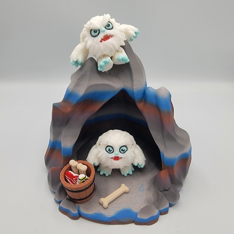 May include: A whimsical, handcrafted diorama featuring two white, furry yeti figures with blue accents and red mouths. The scene includes a cave, a wooden bucket with food, and a bone. The cave is painted in shades of gray, blue, and brown.
