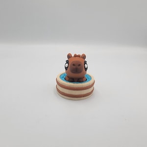 May include: A brown capybara figurine with large eyes sits in a small, round tub. The tub has a light blue interior and is surrounded by a beige and brown exterior. The figurine is centered against a white background.