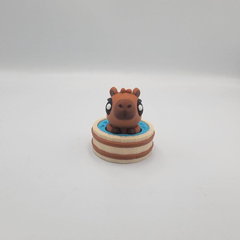 May include: A brown capybara figurine with large eyes sits in a small, round tub. The tub has a light blue interior and is surrounded by a beige and brown exterior. The figurine is centered against a white background.