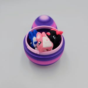 K&ouml;nnte beinhalten: Ein gestreiftes Osterei in Lila und Rosa mit offenem Deckel, in dem sich drei kleine Hasenfiguren befinden. Die Hasen sind rosa, blau und schwarz.