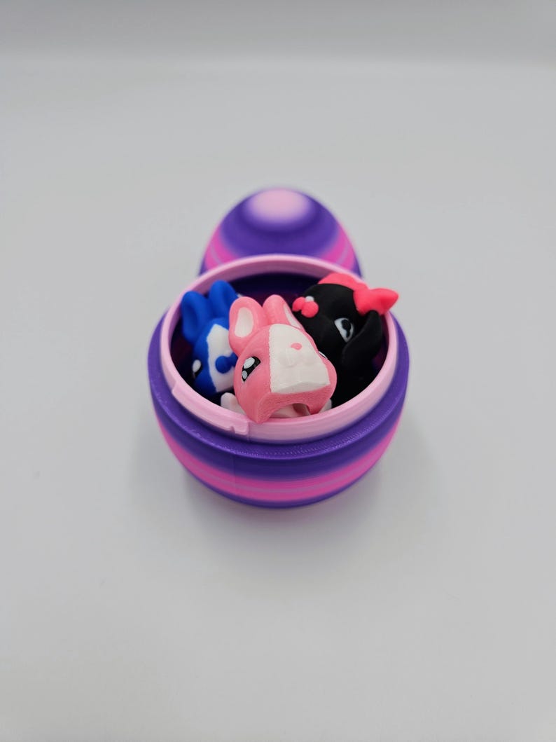 K&ouml;nnte beinhalten: Ein gestreiftes Osterei in Lila und Rosa mit offenem Deckel, in dem sich drei kleine Hasenfiguren befinden. Die Hasen sind rosa, blau und schwarz.