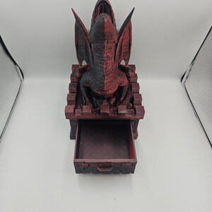 May include: A red and black dragon-themed dice tower with an open drawer. The dragon has its mouth open and wings spread. The tower is made of a dark red material with a textured appearance.