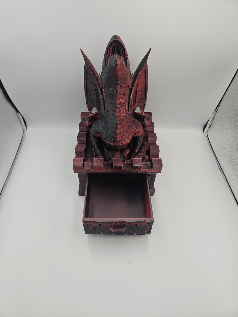 May include: A red and black dragon-themed dice tower with an open drawer. The dragon has its mouth open and wings spread. The tower is made of a dark red material with a textured appearance.