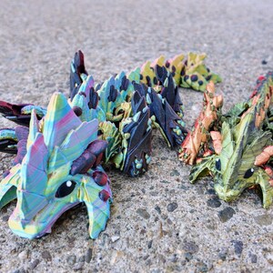May include: Three small, colorful, 3D printed dragon figurines. The dragons are in a line, with the first dragon facing the viewer. The dragon's scales are iridescent and have a variety of colors, including blue, green, purple, and pink. The dragons have black eyes and small, pointed teeth.