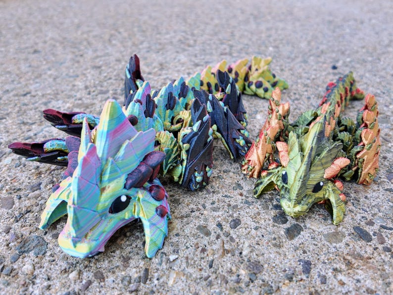 May include: Three small, colorful, 3D printed dragon figurines. The dragons are in a line, with the first dragon facing the viewer. The dragon's scales are iridescent and have a variety of colors, including blue, green, purple, and pink. The dragons have black eyes and small, pointed teeth.