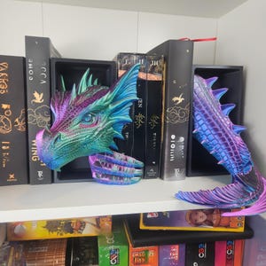 May include: A multi-colored dragon head and tail bookends. The dragon head is a mix of blue, green, purple, and pink hues. The tail is a gradient of purple and blue. The bookends are placed on a white shelf with books.