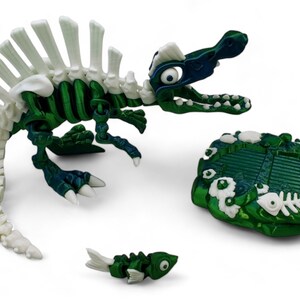 May include: A green and white plastic dinosaur skeleton toy with a white spine and a green body. The dinosaur is standing on a green and white base with a fish bone design. A smaller green and white fish skeleton toy is in the foreground.
