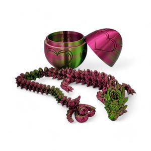May include: A green and pink dragon figurine with a heart design. The dragon is made up of multiple connected pieces and is laying on a white surface. The dragon is in a curled up position with its head resting on its tail. The dragon is a decorative piece and is not a toy.
