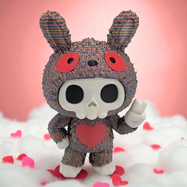 K&ouml;nnte beinhalten: Eine einzigartige Figur eines Hasen mit Totenkopfmaske und Herz auf der Brust. Der Hase ist haupts&auml;chlich grau mit roten Akzenten und wei&szlig;en H&auml;nden und F&uuml;&szlig;en. Er steht auf einem Bett aus wei&szlig;em Flaum mit roten und rosa herzf&ouml;rmigen Konfetti.