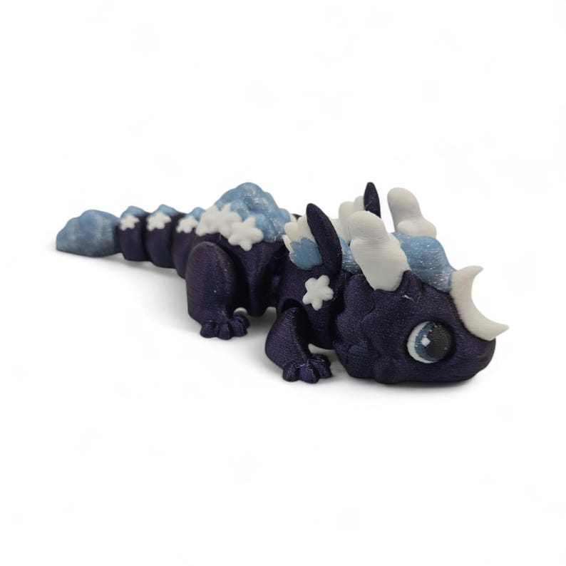 May include: A detailed, 3D-printed dragon figurine in shades of dark blue, light blue, and white. The dragon has a mountain-like structure on its back, snowflake accents, and a crescent-shaped horn. The dragon's eye is a large, detailed, and round blue eye.