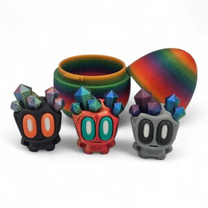 May include: Three colourful, stylised figures with crystal-like tops and large, oval eyes. The figures are black, red, and grey, each with a different colour scheme for the eyes and the numbers "00" on their faces. A matching rainbow-coloured container is in the background.