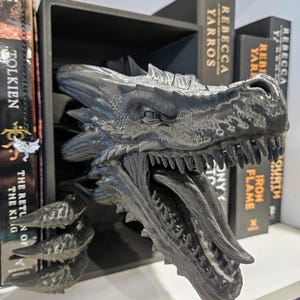 Pode incluir: Uma estante de livros com cabe&ccedil;a de drag&atilde;o preta. A cabe&ccedil;a do drag&atilde;o &eacute; detalhada com escamas e dentes, e est&aacute; posicionada para parecer que est&aacute; a emergir da prateleira. Os livros s&atilde;o vis&iacute;veis na prateleira, com t&iacute;tulos como "Rebecca Yarros".