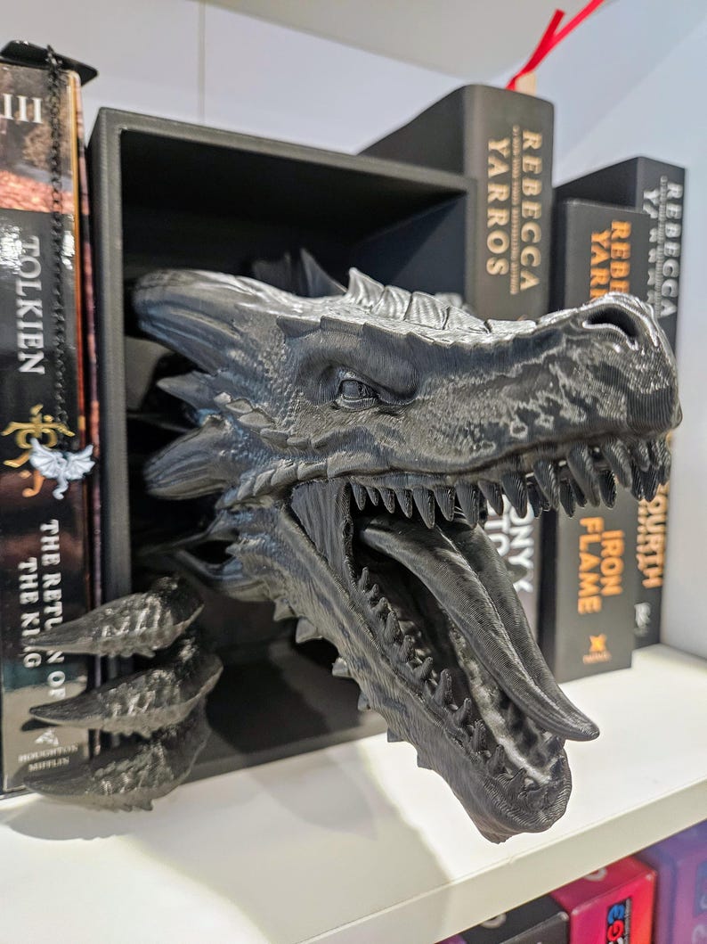 Pode incluir: Uma estante de livros com cabe&ccedil;a de drag&atilde;o preta. A cabe&ccedil;a do drag&atilde;o &eacute; detalhada com escamas e dentes, e est&aacute; posicionada para parecer que est&aacute; a emergir da prateleira. Os livros s&atilde;o vis&iacute;veis na prateleira, com t&iacute;tulos como "Rebecca Yarros".