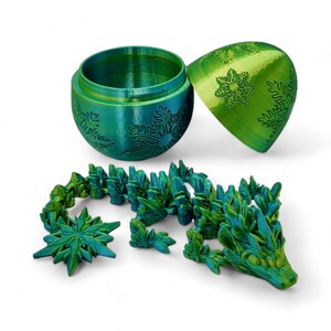 K&ouml;nnte beinhalten: Ein gr&uuml;ner und blauer 3D-gedruckter Weihnachtsbaumschmuck in Form eines Eies mit einem Schneeflockenmuster. Der Schmuck ist ge&ouml;ffnet und ein gr&uuml;ner und blauer 3D-gedruckter Drache liegt auf dem Tisch.