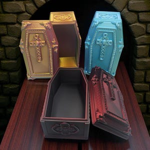 May include: Four decorative coffin-shaped boxes in various colors: gold, pink, blue, and red. Each coffin features a cross design and ornate detailing. The boxes are open or partially open, revealing their interiors. They are arranged on a dark wood surface.