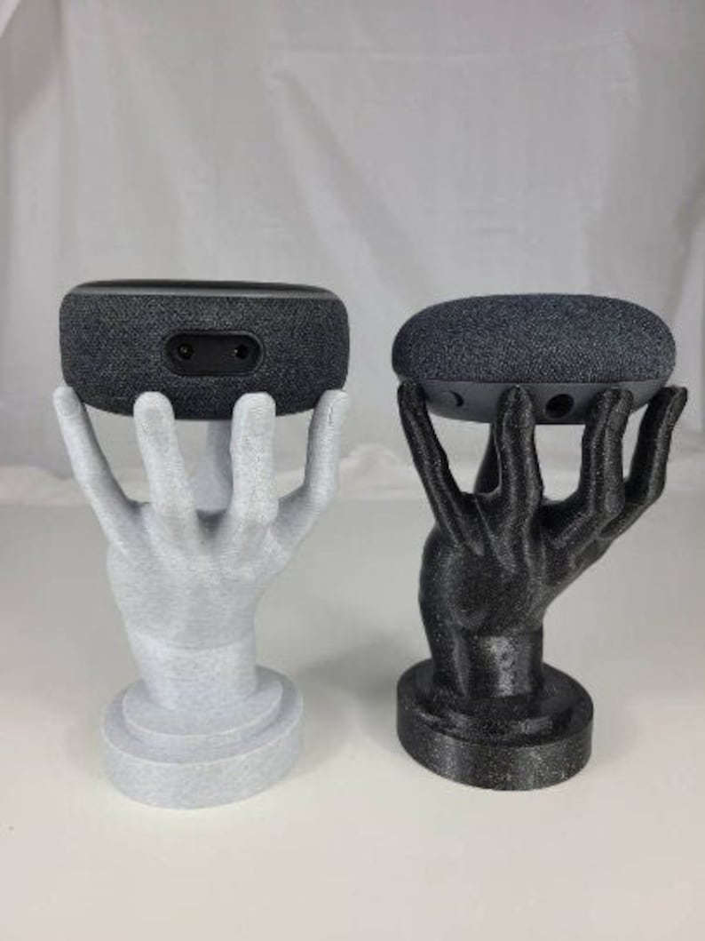 Op de afbeelding: Twee 3D-geprinte handen die een zwarte en een grijze slimme speaker vasthouden. De handen zijn in lichtgrijs en zwart geverfd.