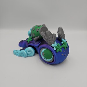 Pode incluir: Uma escultura colorida e fantasiosa de uma criatura com um olho grande e detalhes florais. A escultura apresenta um gradiente de azul e roxo, com detalhes verdes e cinzentos. A criatura tem uma cabe&ccedil;a cil&iacute;ndrica com um topo verde e um design floral estilizado.