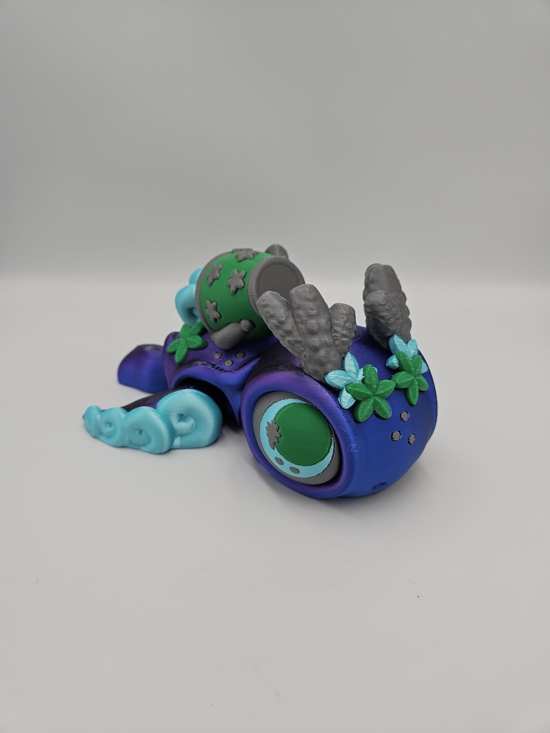 Pode incluir: Uma escultura colorida e fantasiosa de uma criatura com um olho grande e detalhes florais. A escultura apresenta um gradiente de azul e roxo, com detalhes verdes e cinzentos. A criatura tem uma cabe&ccedil;a cil&iacute;ndrica com um topo verde e um design floral estilizado.