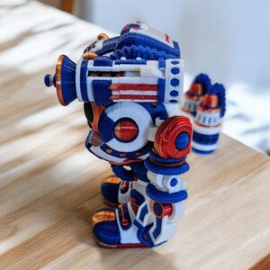 May include: A 3D printed robot toy in blue, white and red. The robot has a round body with a large head and two arms. The robot is standing on a wooden surface.