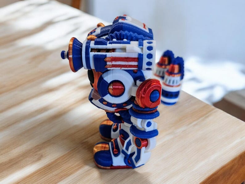 May include: A 3D printed robot toy in blue, white and red. The robot has a round body with a large head and two arms. The robot is standing on a wooden surface.
