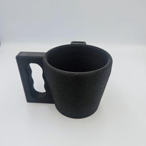 Pu&ograve; includere: Una tazza nera con una superficie strutturata e un'ampia maniglia rettangolare. Il manico ha un ritaglio per l'impugnatura. La tazza &egrave; cilindrica con una parte superiore leggermente svasata.